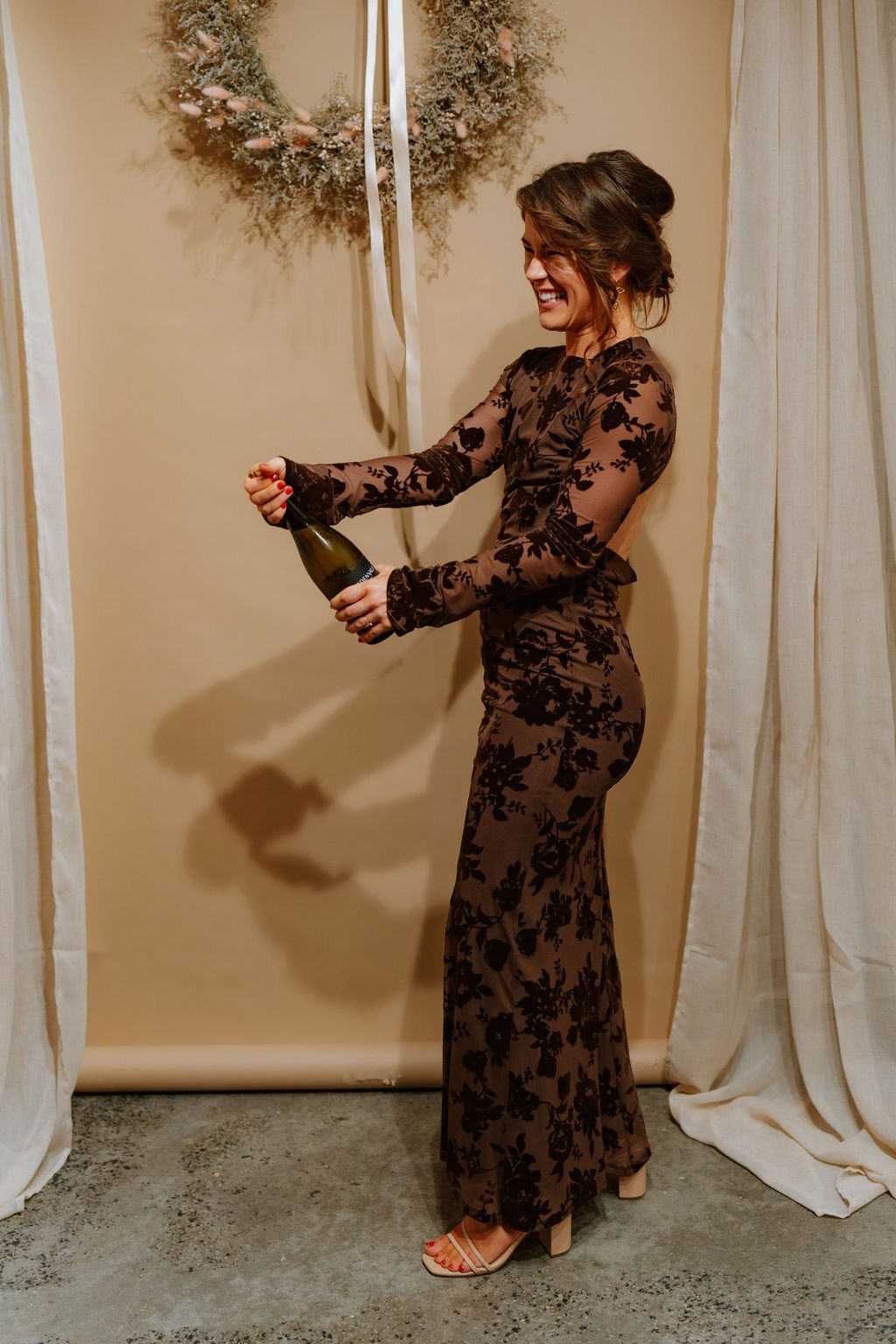 A woman in a sheer, long-sleeved, floral jumpsuit opens a bottle of champagne with a big smile.