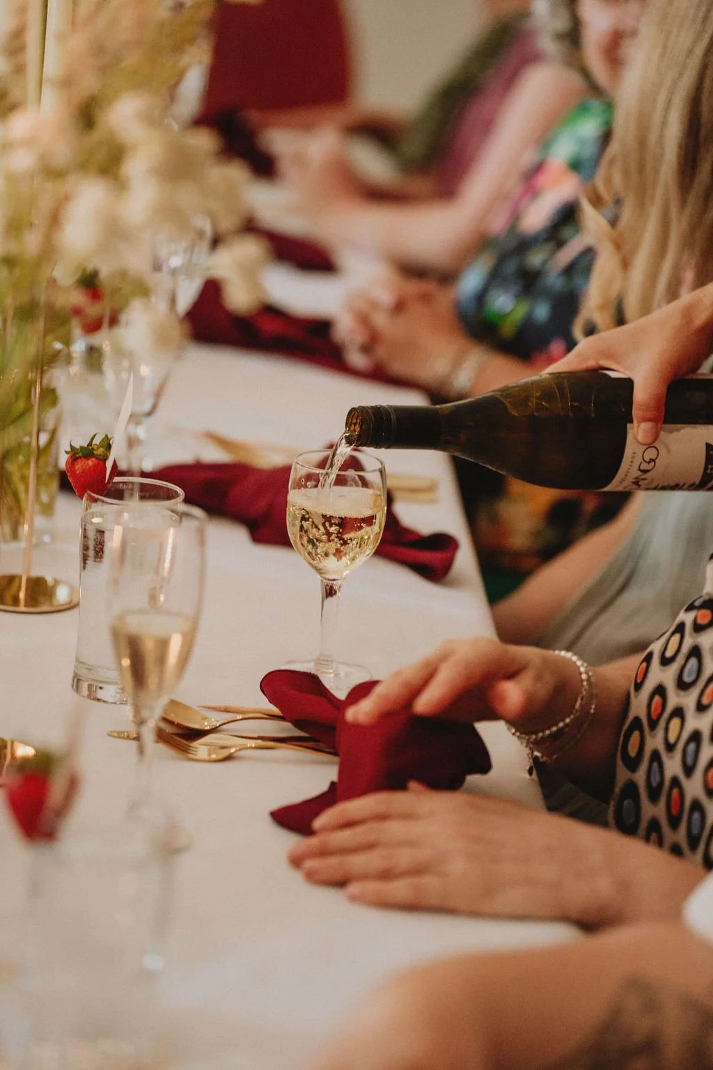 A person pouring champagne into a flute at a formal gathering with other guests seated at a decorated table.