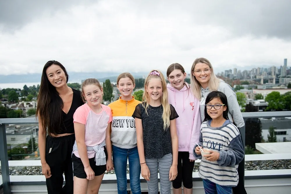 Group of seven smiling young girls and women standing outdoors on a rooftop with a cityscape and cloudy sky in the background.