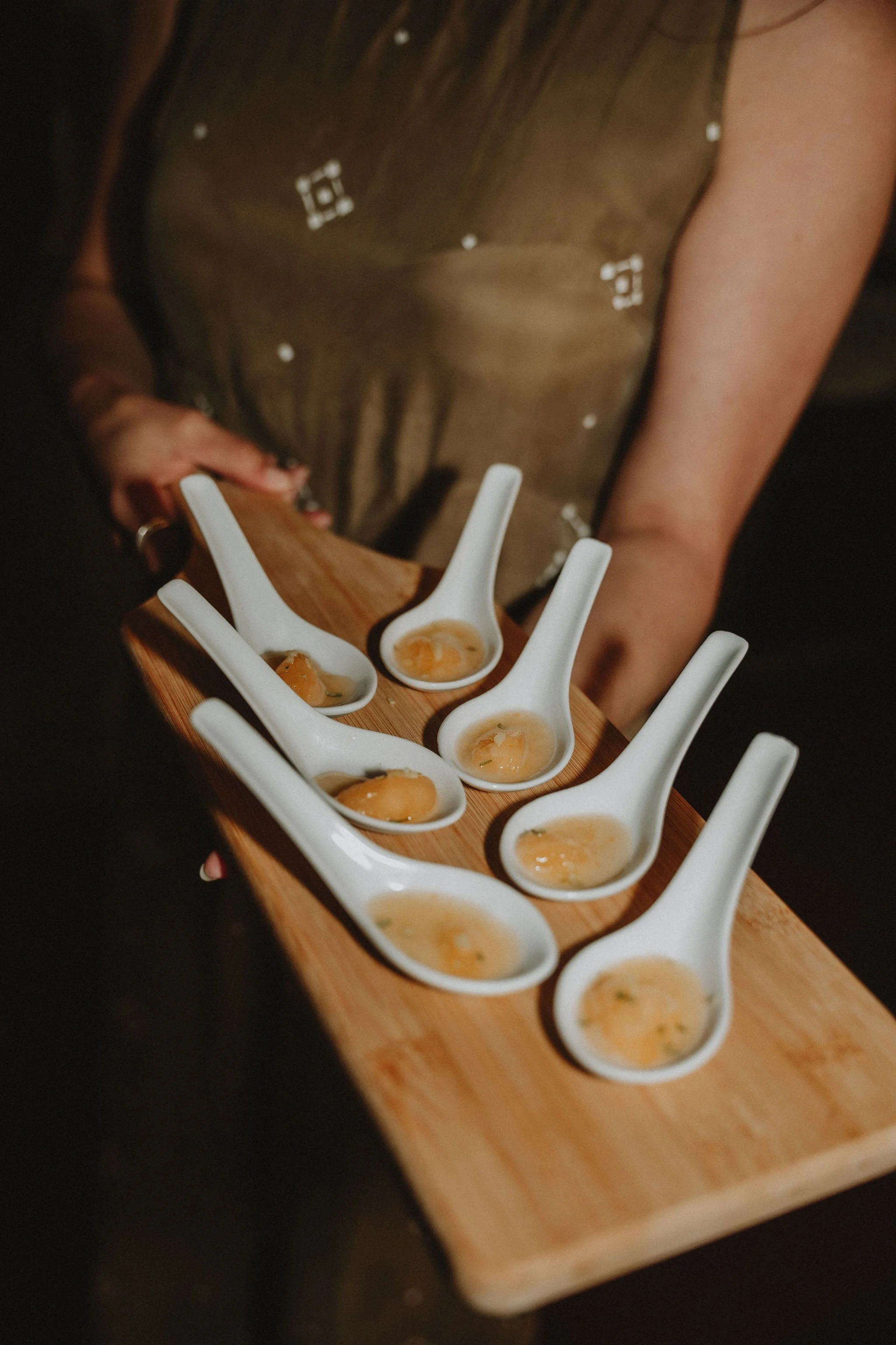 A person holding a wooden tray with six white ceramic spoons containing small servings of a food, possibly egg-based, in a dimly lit setting.