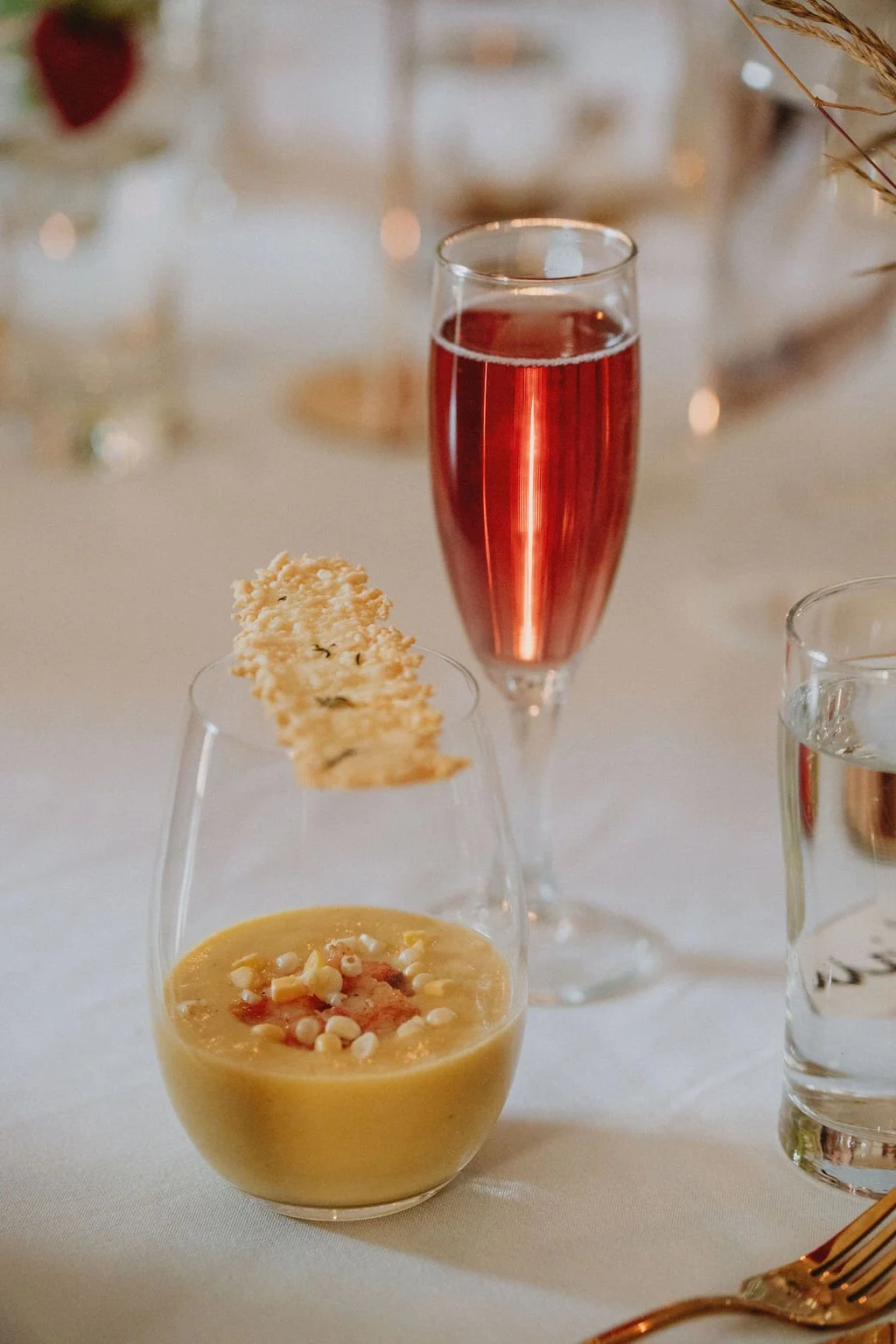 A glass of pink sparkling wine, a small bowl of creamy yellow soup with toppings, and a glass of water on a table.