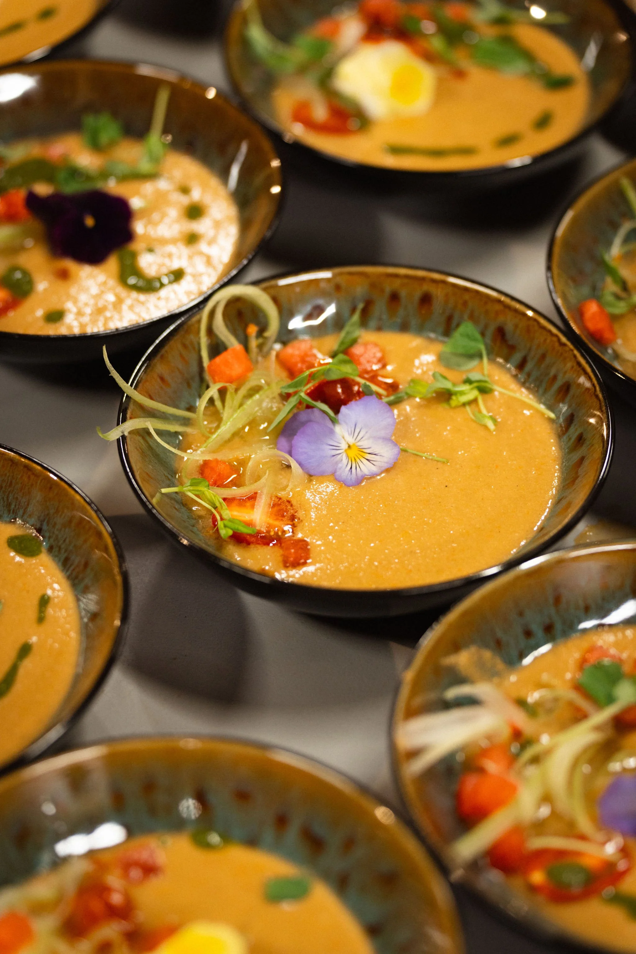 Bowls of colorful soup garnished with edible flowers, vegetables, and herbs.