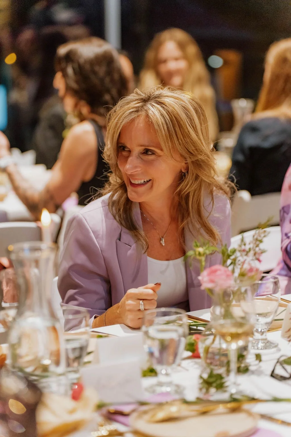 A woman with shoulder-length blonde hair, wearing a lavender blazer and white top, smiling and engaged in conversation at a dinner table decorated with pink flowers and glassware, at a social gathering or celebration.