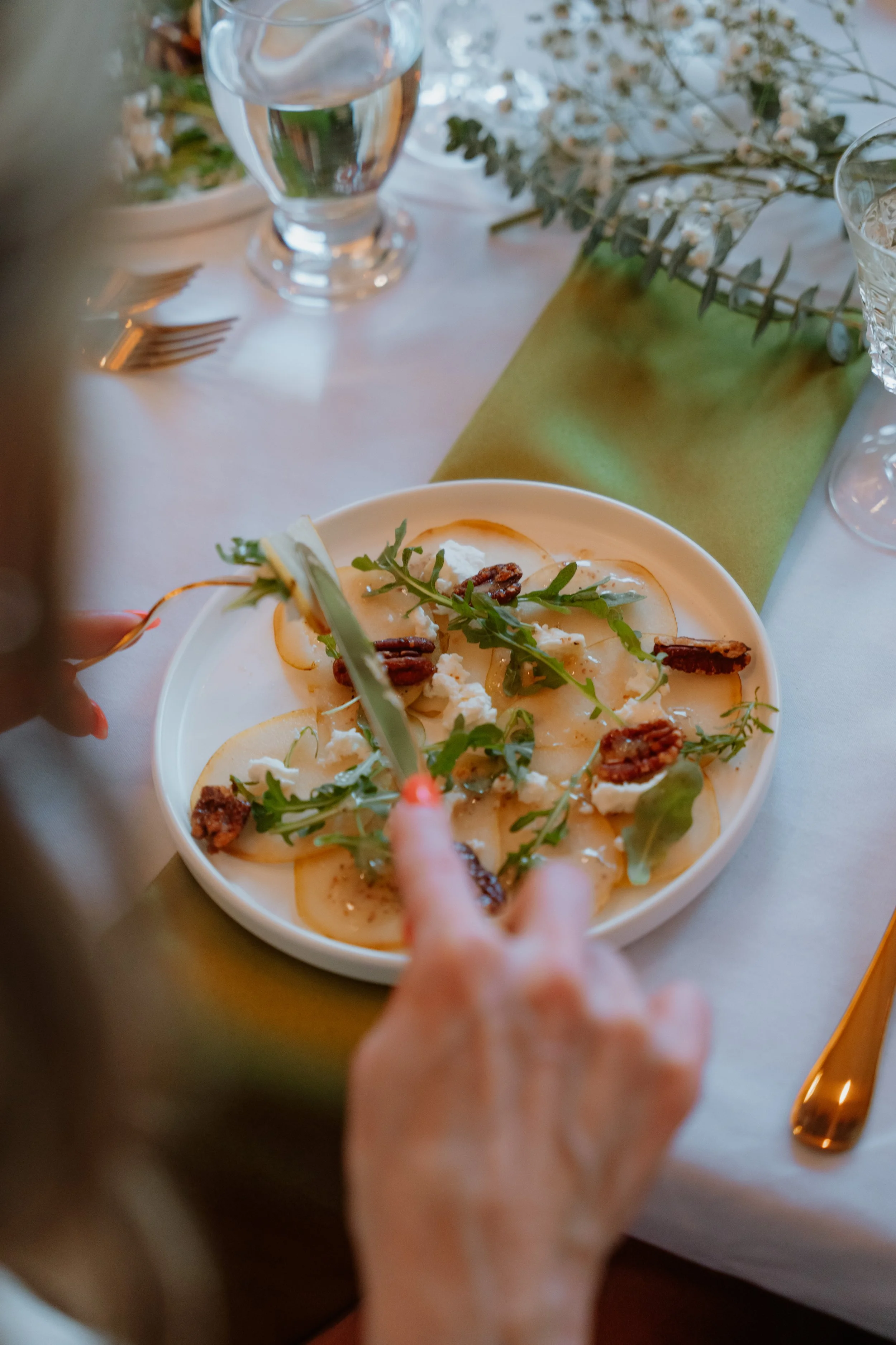 A person is serving themselves a plate of cheese, pear slices, arugula, and pecans at a dining table set with glassware, utensils, and a green napkin.