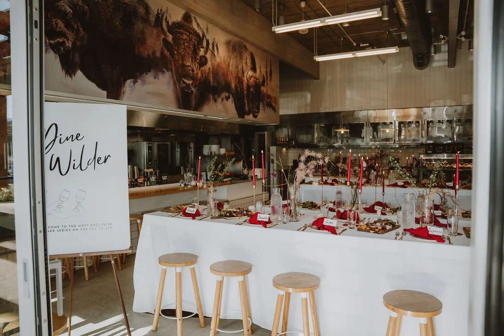 Interior of a restaurant or event space with a long table set with red napkins, candles, and place cards. Behind the table, there is a mural of bison on the wall. A sign on an easel reads "Jinne Wilder".