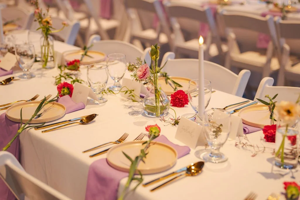 Wedding reception table decorated with pink and red flowers, gold cutlery, purple napkins, glasses, and a lit white candle in the center.