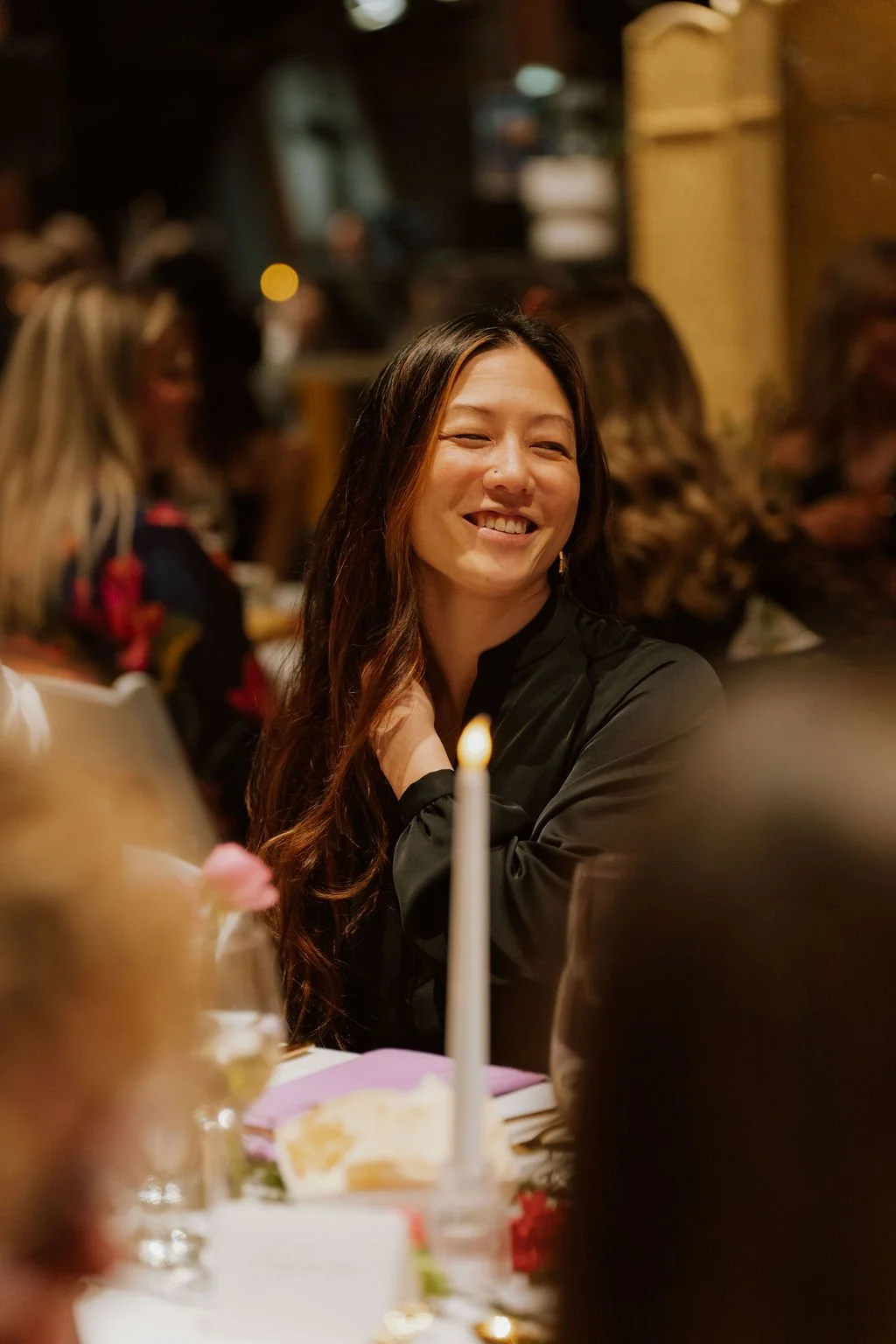 A woman with long dark hair and a black top smiling in a dimly lit social setting, with blurred people in the background and a lit candle on a decorated table in the foreground.