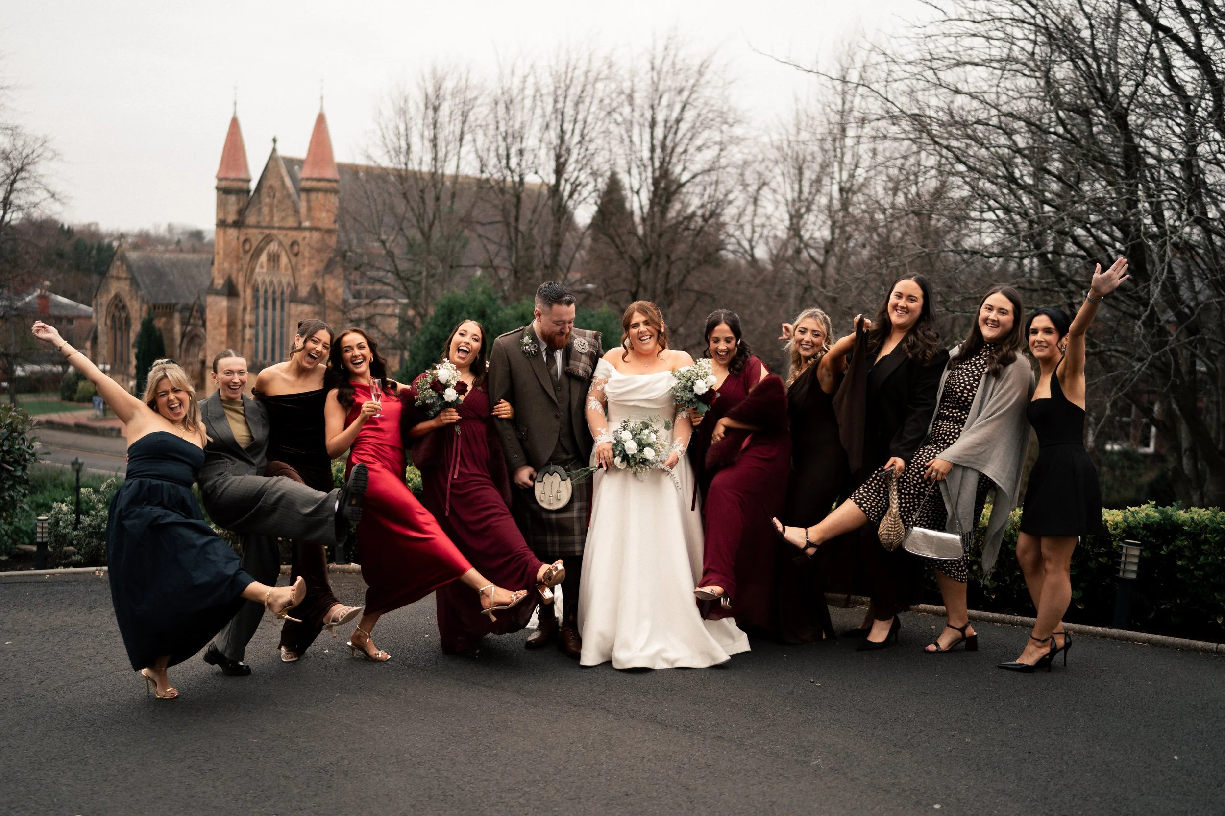 Group of people celebrating at a wedding, standing outdoors on a paved area with a church in the background, some holding bouquets and wine glasses, wearing formal attire, and smiling joyfully.