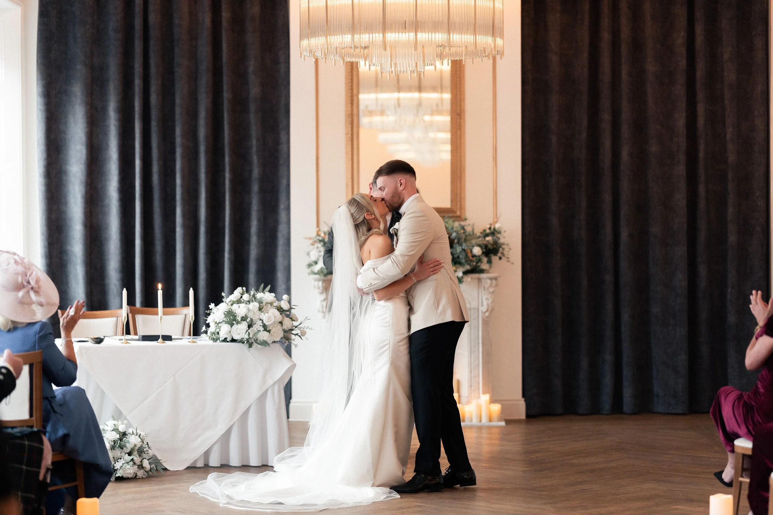 A bride and groom share a kiss during their wedding ceremony in a decorated indoor venue with candles and floral arrangements.