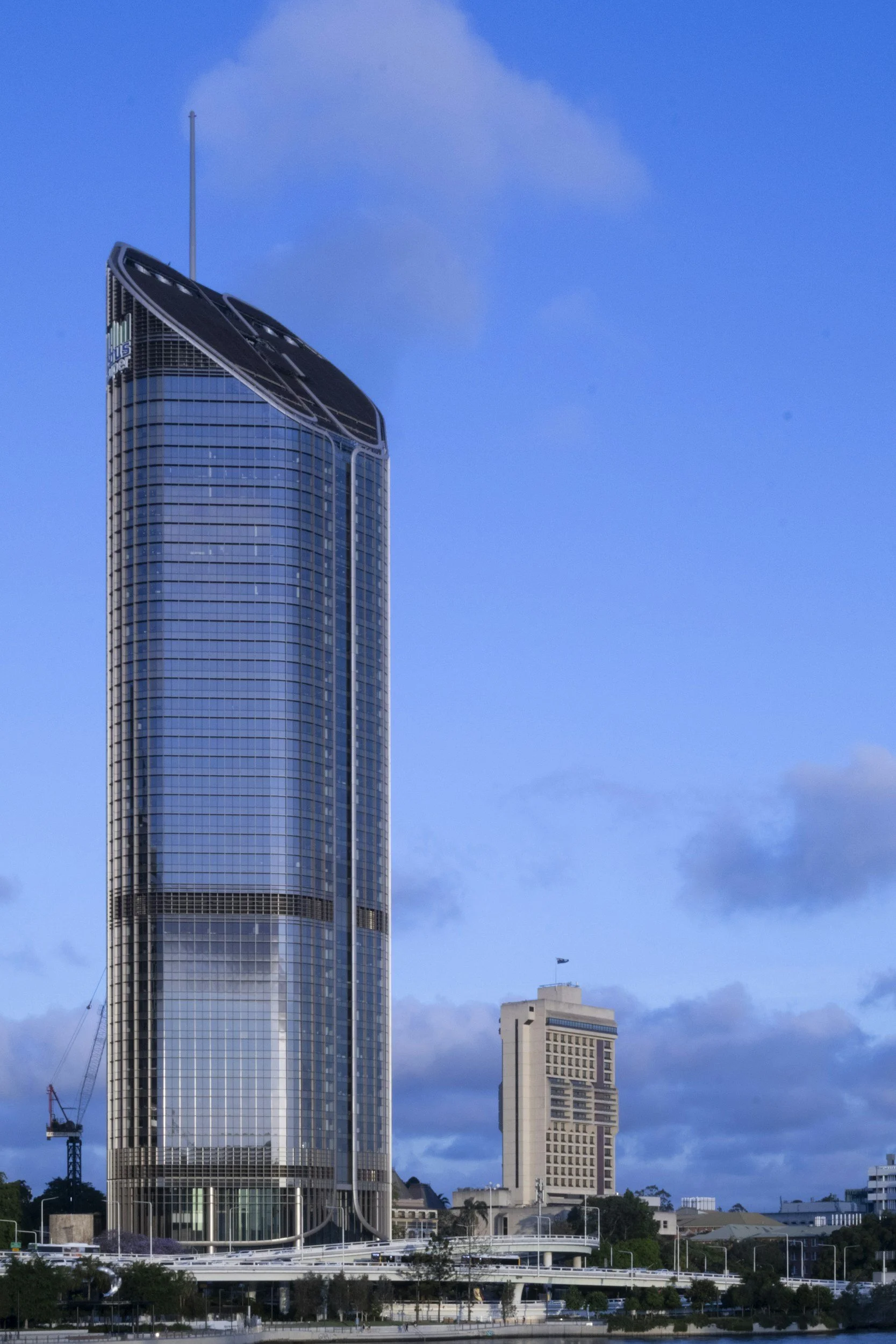 A modern skyscraper with a curved top and glass facade, situated next to a shorter building, under a blue sky with some clouds.