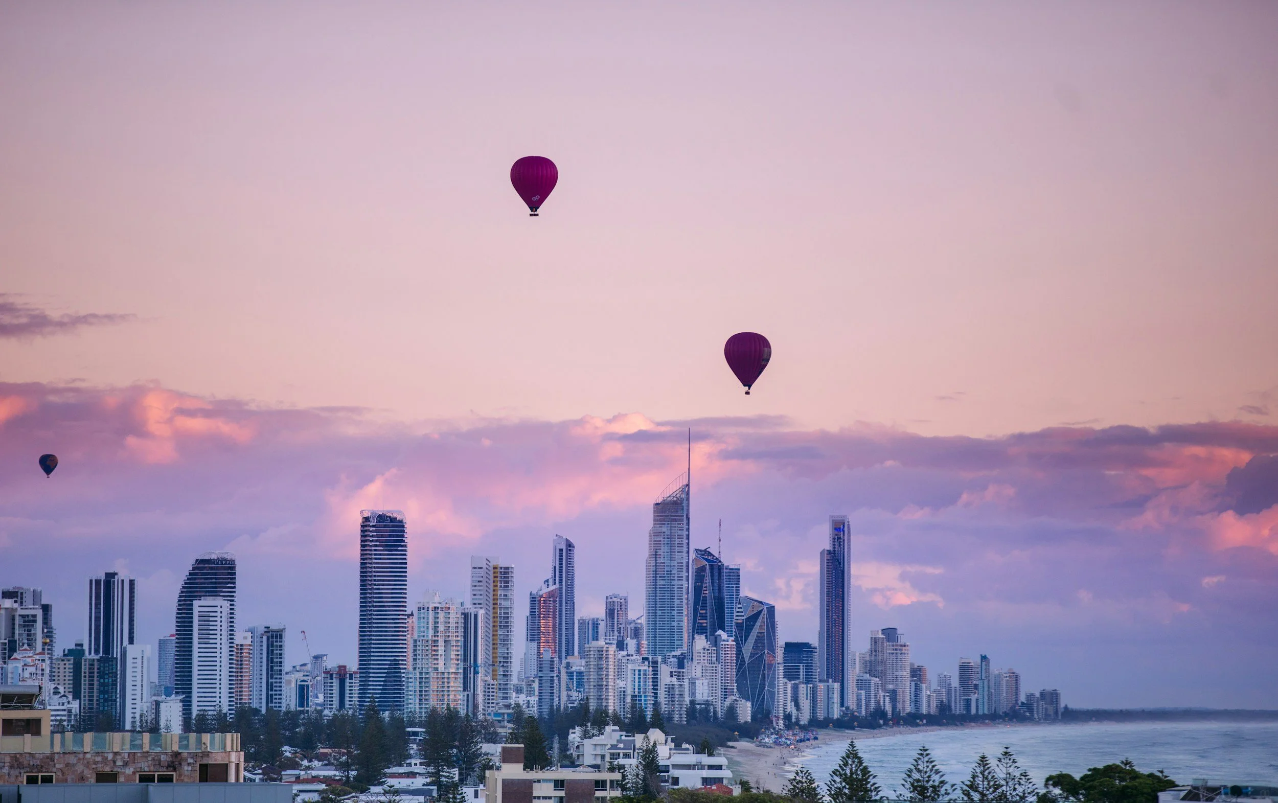 Skyline view of a city with high-rise buildings and three hot air balloons floating in the sky during sunset or dawn.