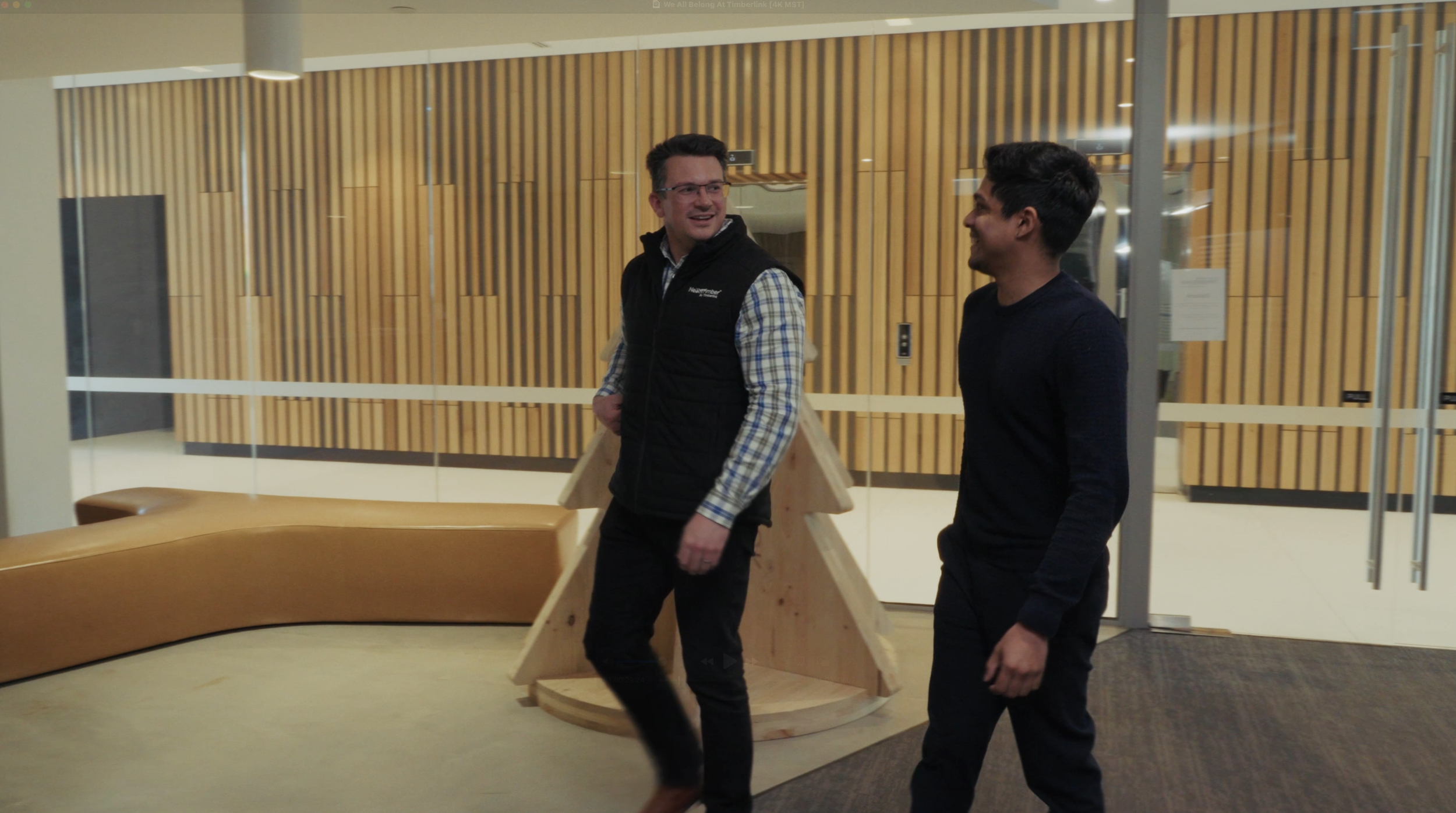 Two men smiling and walking in a modern indoor space with wooden wall decor and glass doors, engaged in conversation.