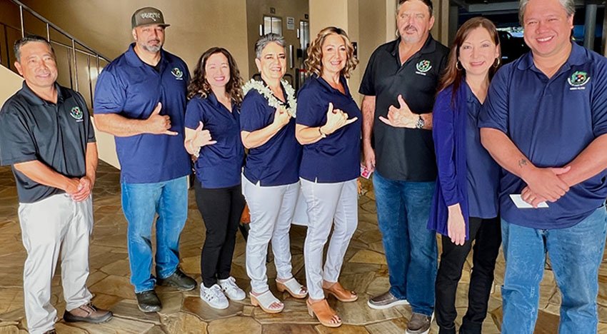 A group of people standing and posing for a photo indoors, all wearing blue shirts, with some making shaka hand signs.
