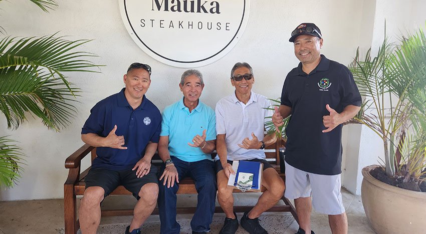 Four men posing with shaka hand signs in front of a Maka Steakhouse sign, two sitting on a bench, two standing, surrounded by palm plants.