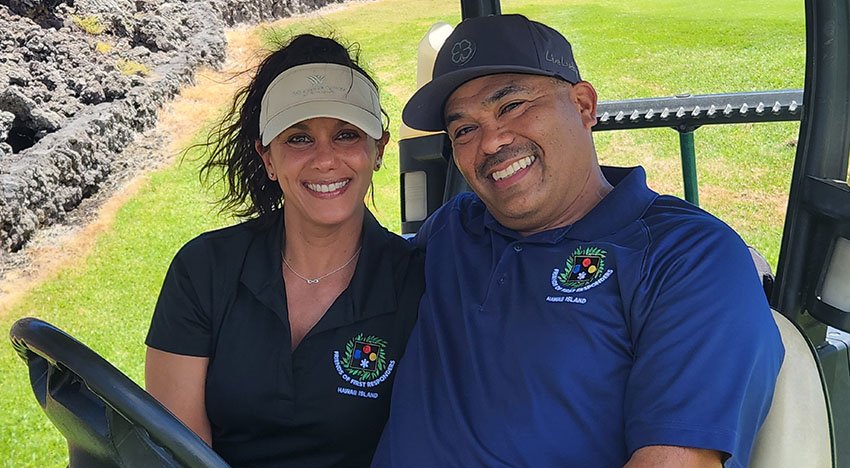 A smiling couple sitting in a golf cart on a grassy area, wearing casual shirts and caps.
