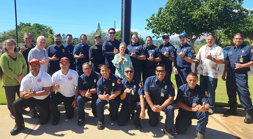 A group of firefighters posing outdoors with a mixture of uniforms and casual clothing, making shaka hand signs.