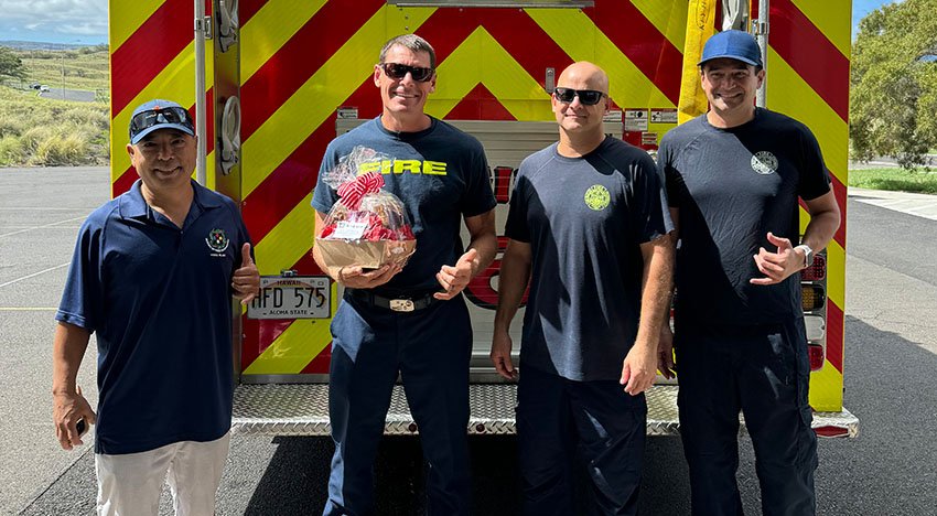 Four firefighters standing in front of a fire truck, one holding a gift basket, all are smiling and giving thumbs up.