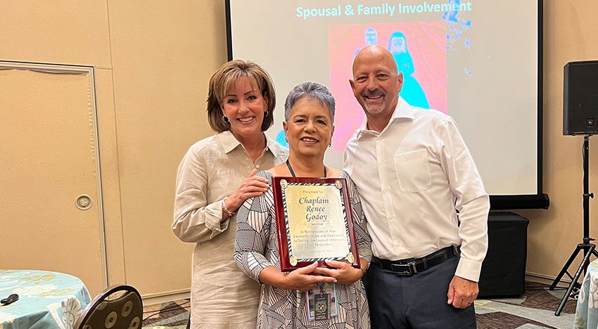 Three people standing together indoors, one holding a framed certificate or plaque. There's a presentation screen in the background.