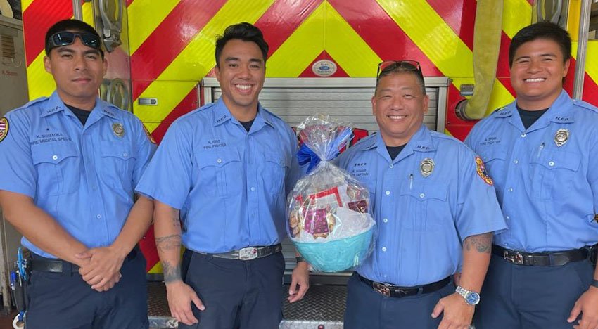 Four firefighters in blue uniforms standing in front of a fire truck holding a gift basket.