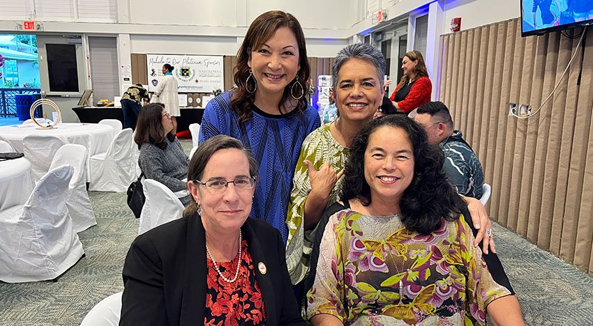 Group of four women smiling indoors at a social gathering with tables and chairs in the background.