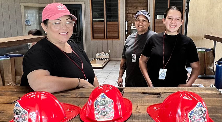 Three people smiling behind a wooden counter, with four red plastic fire helmets in the foreground. They are in a room with wooden walls and windows.