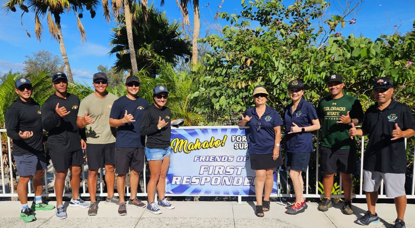 A group of people standing in front of a sign that says "Mahalo! For Your Support Friends of First Responders." They are outdoors in a sunny area with palm trees and greenery in the background. Many are making a "shaka" hand gesture and are casually