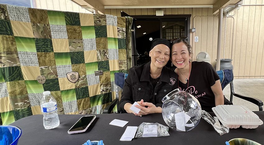 Two women smiling at an outdoor table with a black tablecloth, a quilt in the background, and various items such as a water bottle, smartphone, and a clear bowl with notes and envelopes.