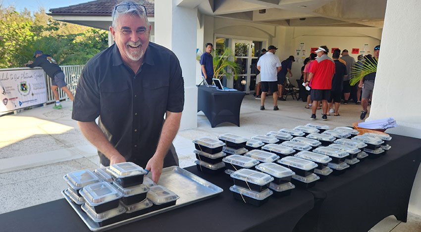 Man smiling while arranging stacked food containers on a table outdoors, with people gathered in the background at an event.