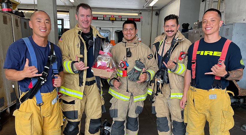 Five firefighters, wearing various parts of their gear, stand together in a fire station or garage. They are smiling and gesturing with their hands. One is holding a gift basket, indicating a celebratory moment.