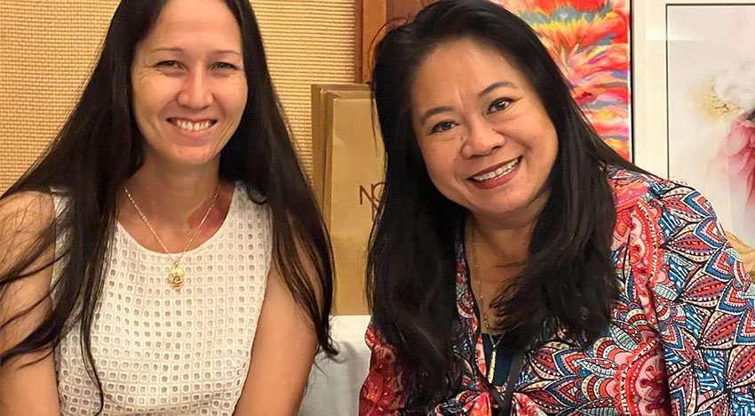 Two women smiling, sitting indoors with colorful decor in the background.