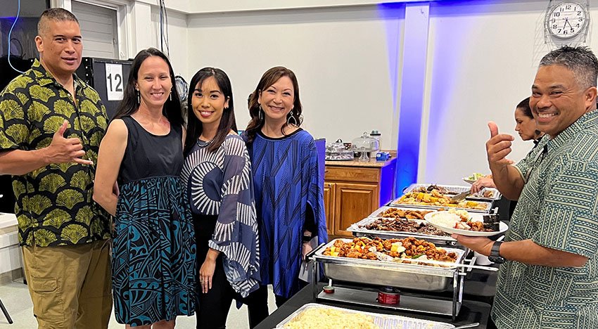Group of five people standing next to a buffet table with various dishes, smiling and posing for a photo indoors. The table is filled with trays of food, and there is a clock on the wall above.