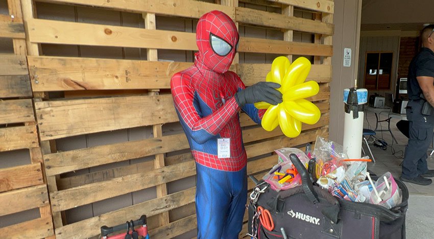Person in Spider-Man costume making a yellow balloon sculpture next to a toolbox against a wooden pallet wall.