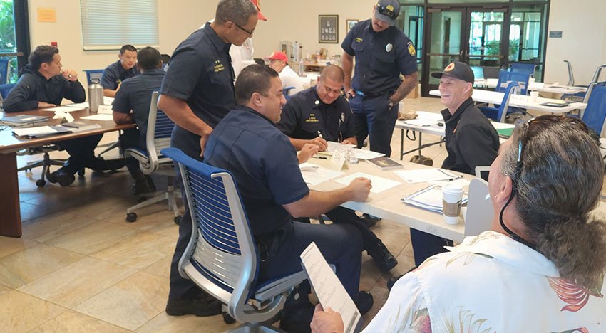 Group of people in navy uniforms sitting and standing around tables in a conference room, engaged in a discussion or training session with documents and coffee cups on tables.