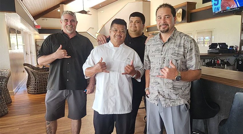 Four men standing in a bar or restaurant setting, all making a shaka hand gesture. One man is wearing a chef's uniform, while the others are in casual shirts. They appear to be posing together, smiling.