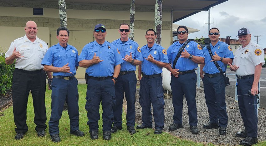 Group of people in blue uniforms standing on grass, with several making a hand gesture. They appear to be part of a public safety or service team, possibly firefighters or EMTs. Two people in white uniforms are also present on either side of the grou