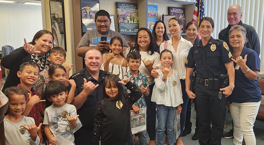 A group of adults and children smiling and gesturing with shaka signs in an office setting. Some are wearing police uniforms, and there are posters and an American flag in the background.
