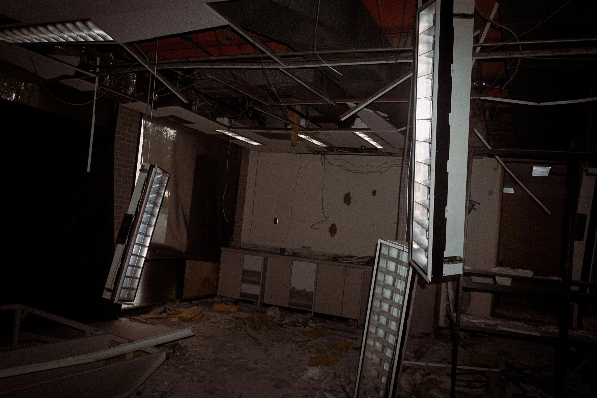 Dark, abandoned room with damaged ceiling, scattered debris on the floor, and broken light fixtures. The room appears to be a kitchen area with some cabinets and pipes visible.