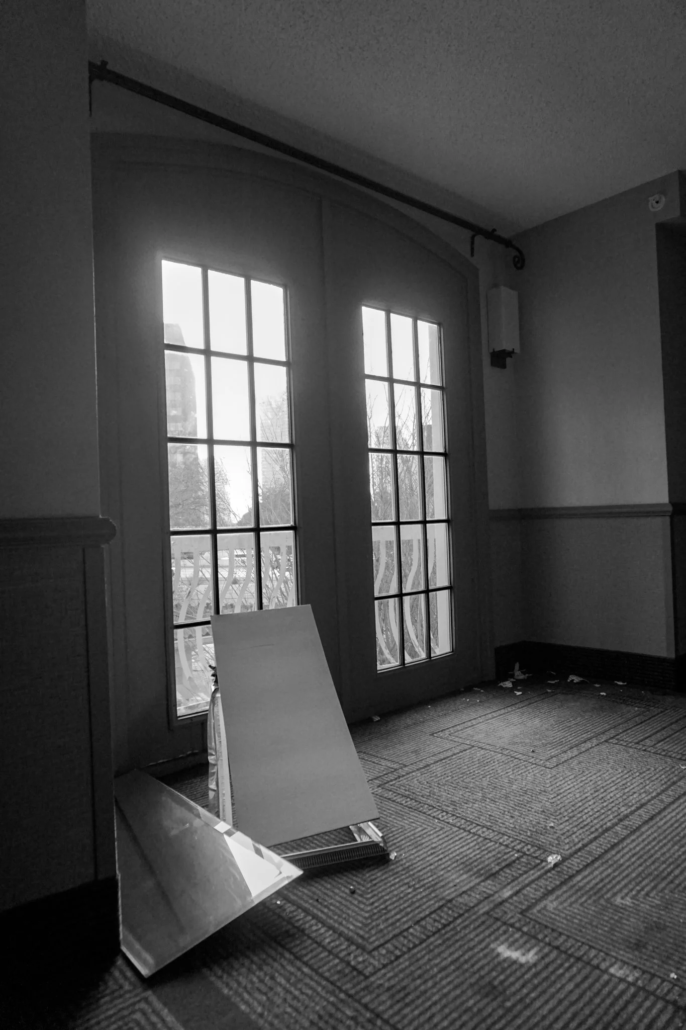 Black and white photo of a room with closed glass double doors, a carpeted floor, and an interior wall with a horizontal wood panel. A mirror and a blank board are leaning against the wall, with small debris scattered on the floor.