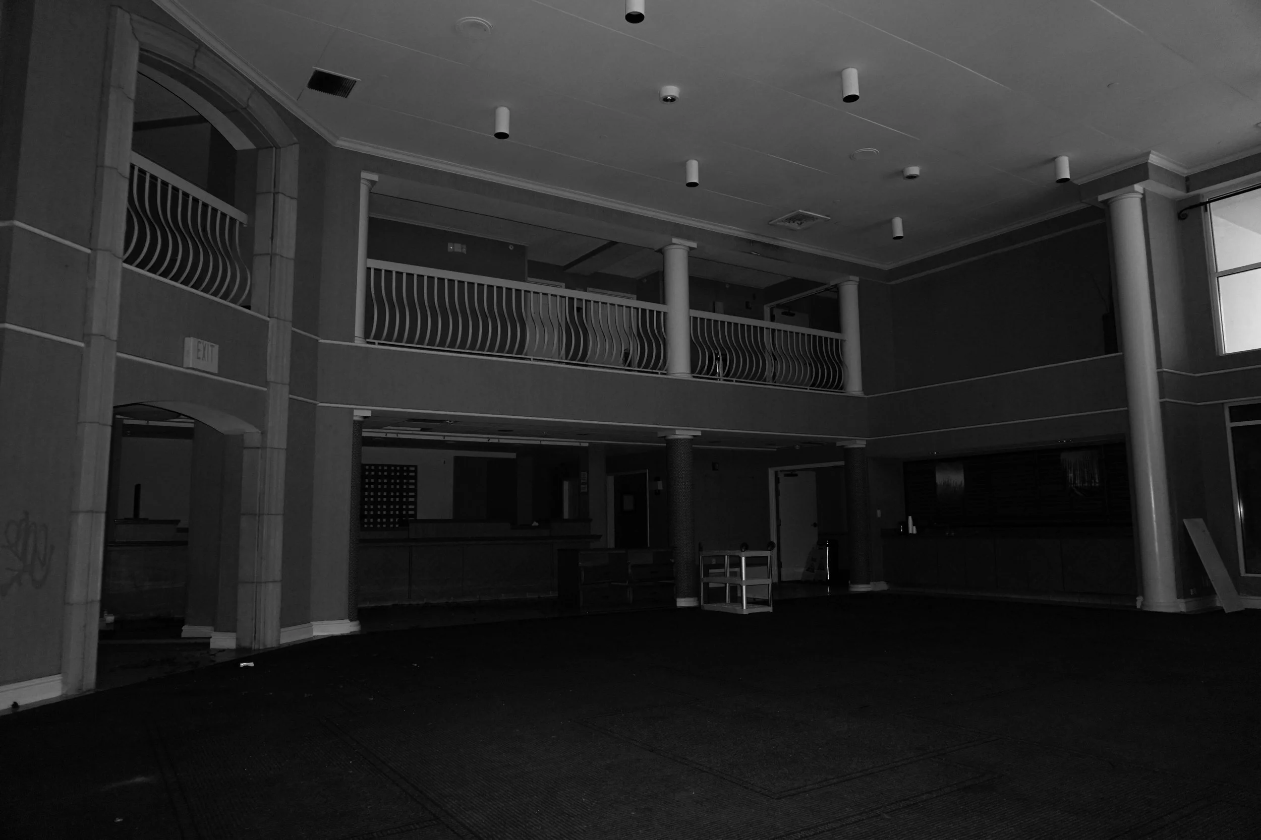 An empty, dimly lit indoor space with a two-story interior, featuring a balcony with curved railings, columns, and large windows.