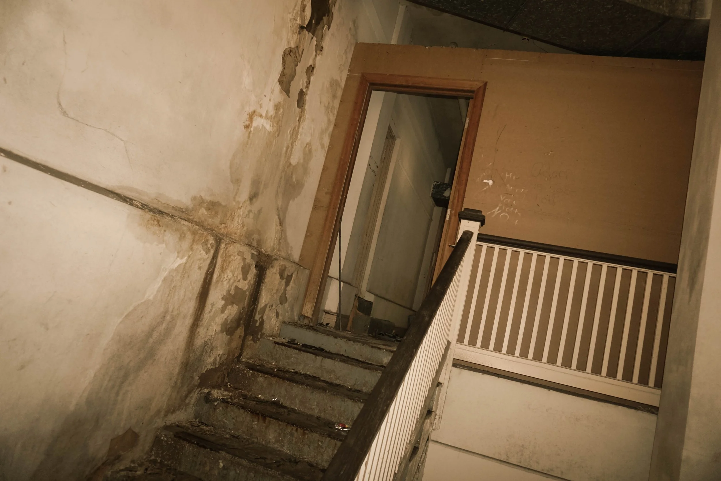 A damaged interior staircase with worn concrete steps leading up to a doorway. The surrounding walls are stained and peeling. There is a wooden frame around the doorway, and a staircase railing with vertical metal bars on the right.