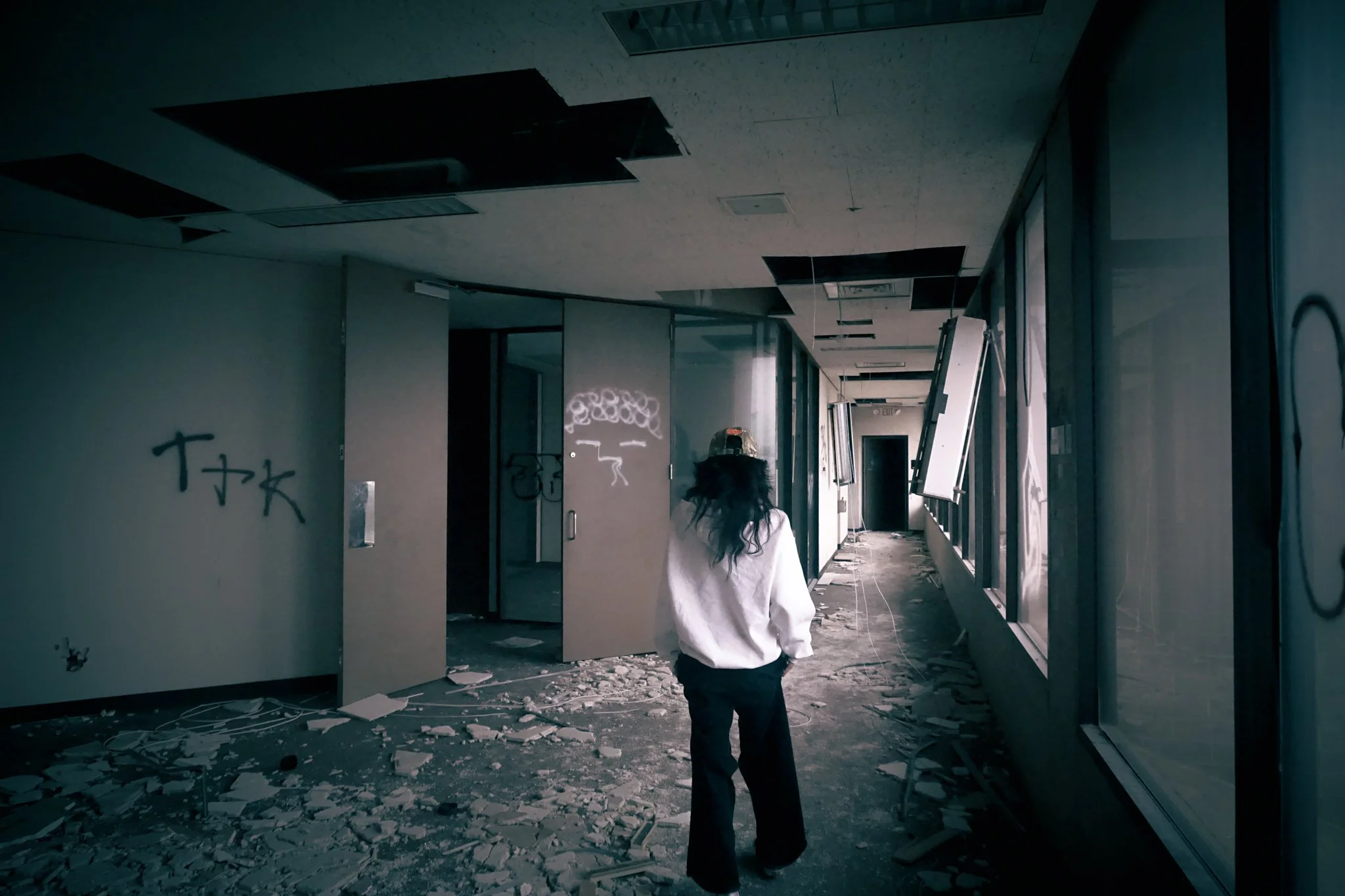 A woman with long dark hair wearing a white sweater and dark pants exploring an abandoned building with debris on the floor, broken ceiling tiles, and graffiti on the walls.