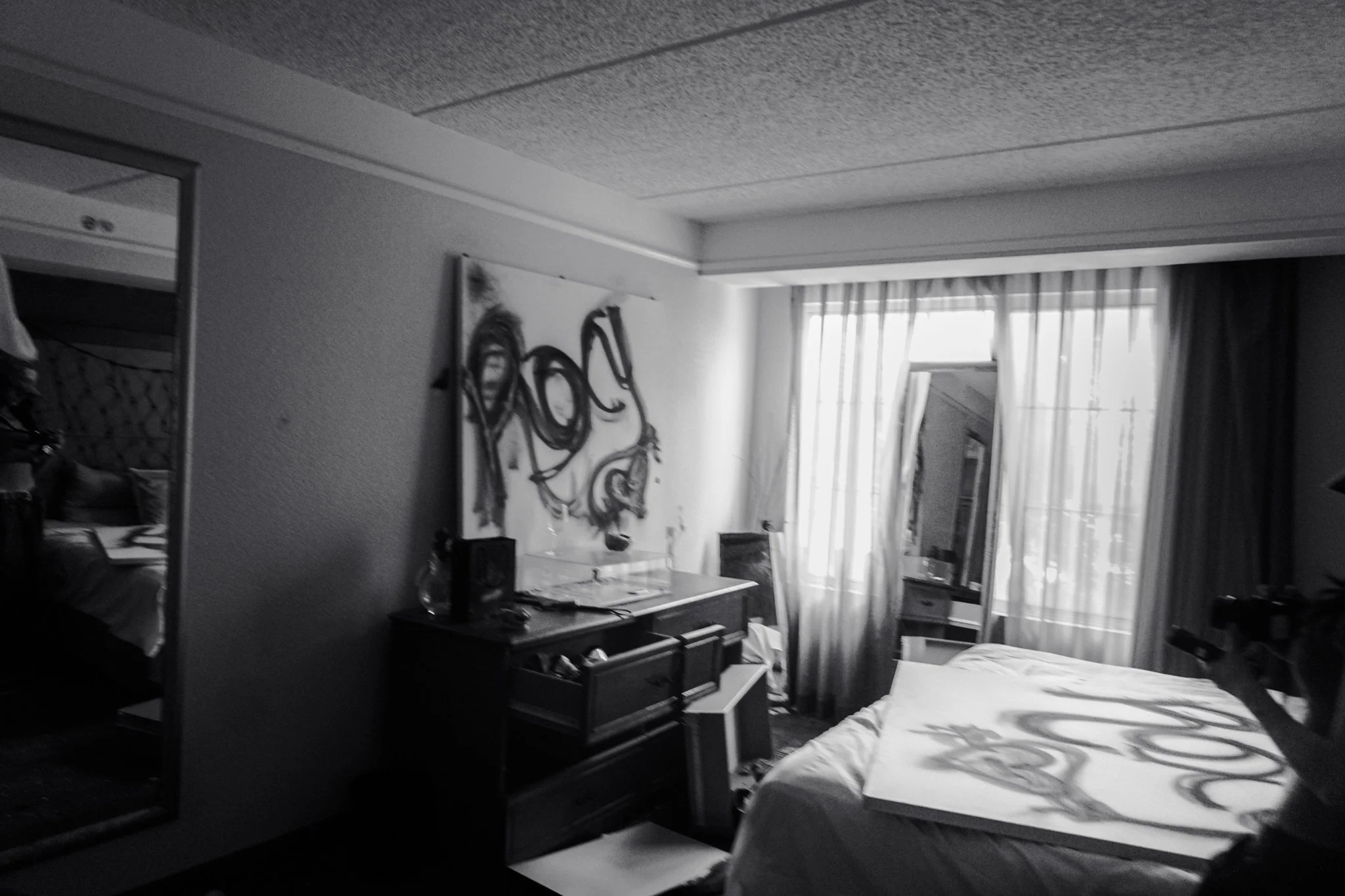 A black and white photo of a hotel room with a bed, dresser, mirror, and a large window with curtains.