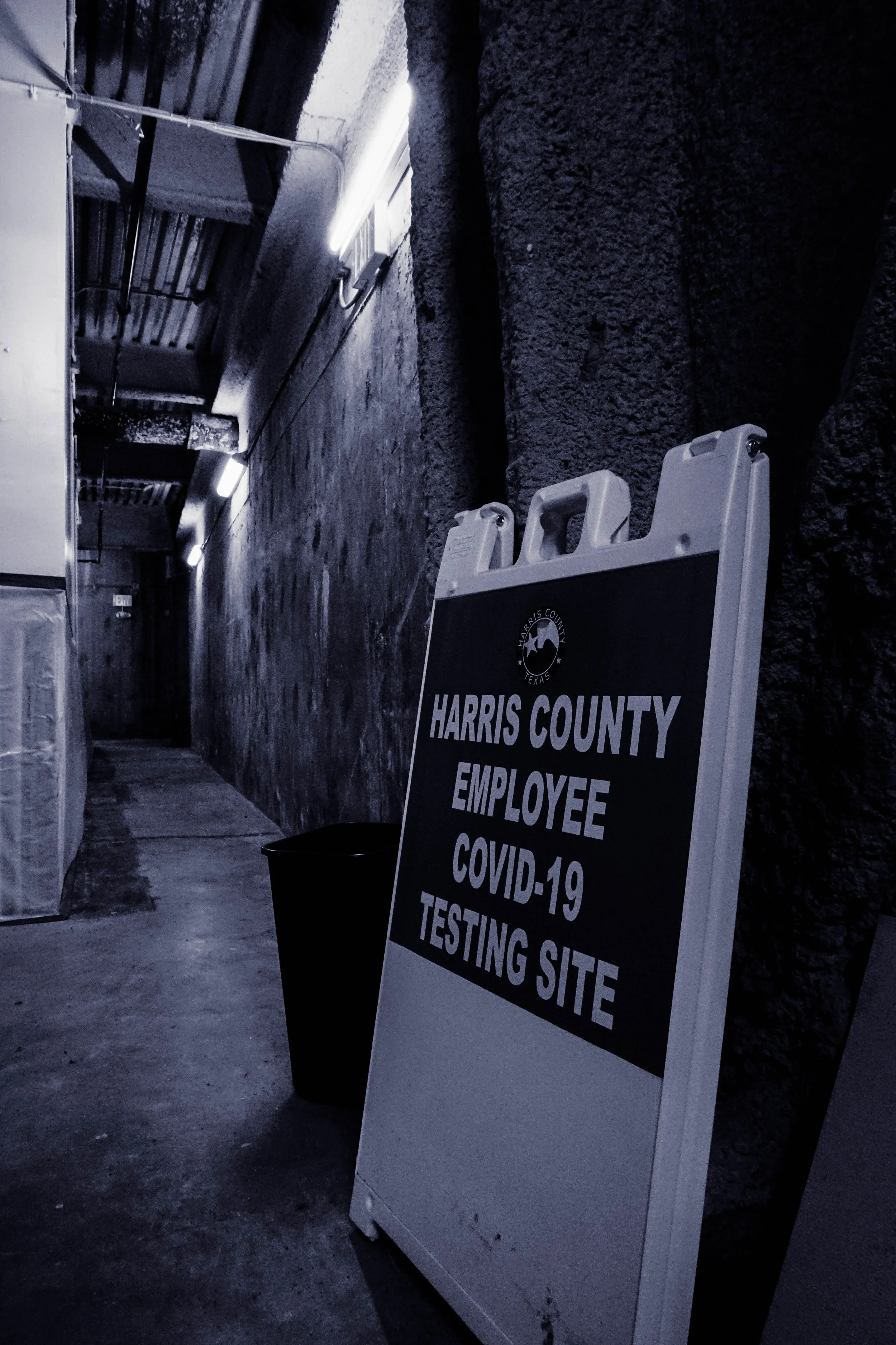 A black and white sign indicating Harris County employee COVID-19 testing site inside a dimly lit corridor with exposed ceiling pipes and textured walls.