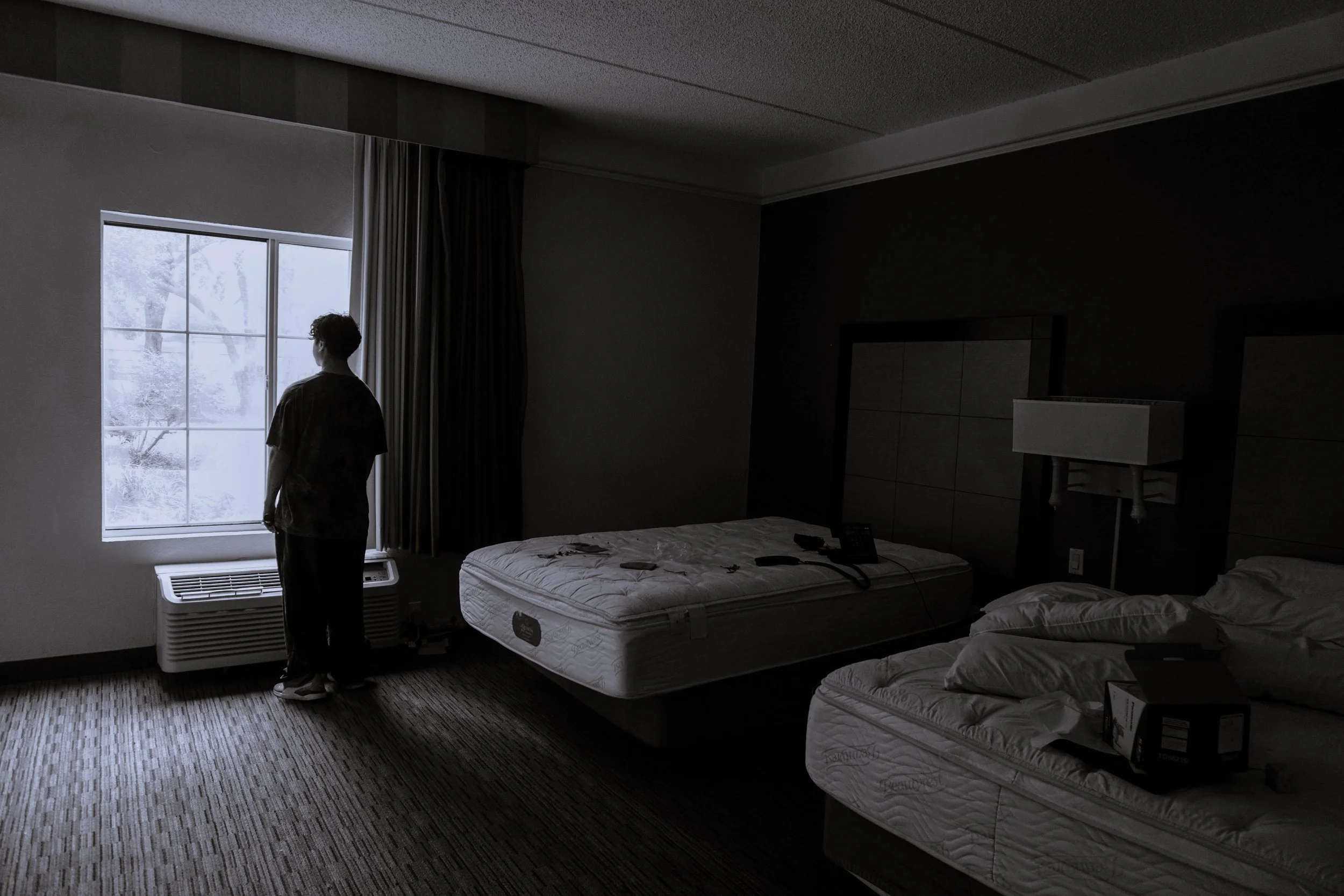 A man standing by a window in a dark hotel room, looking outside at winter snow-covered trees, with two unmade beds, a bedside lamp, and a few items on the beds.