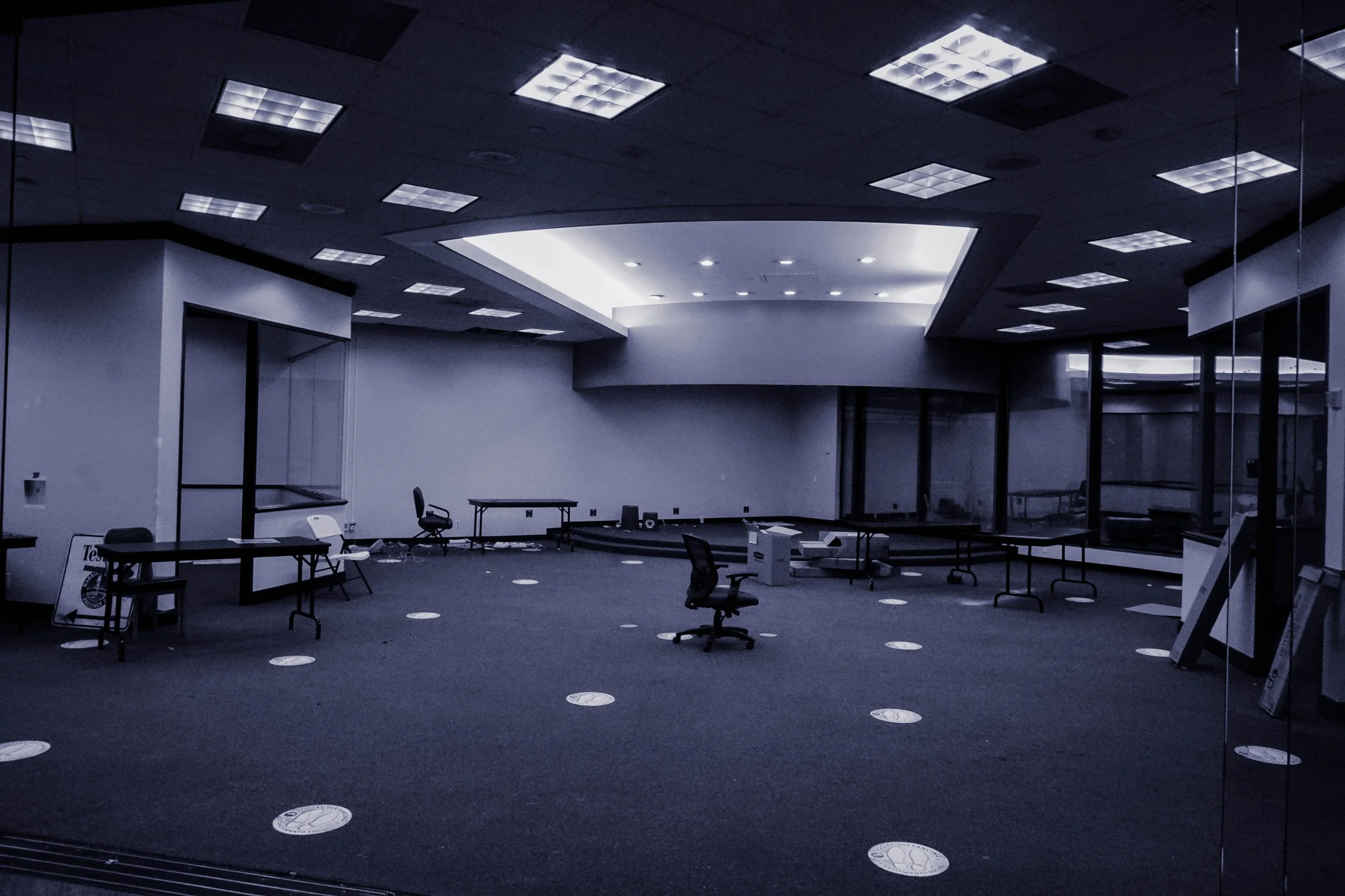 Empty office space with scattered chairs and tables, no people, and social distancing markers on the carpet.