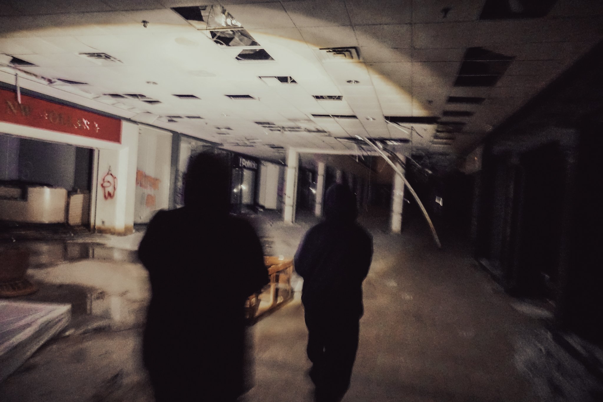 Two silhouetted people walking in an abandoned, damaged shopping mall with a collapsed ceiling, broken tiles, and debris.