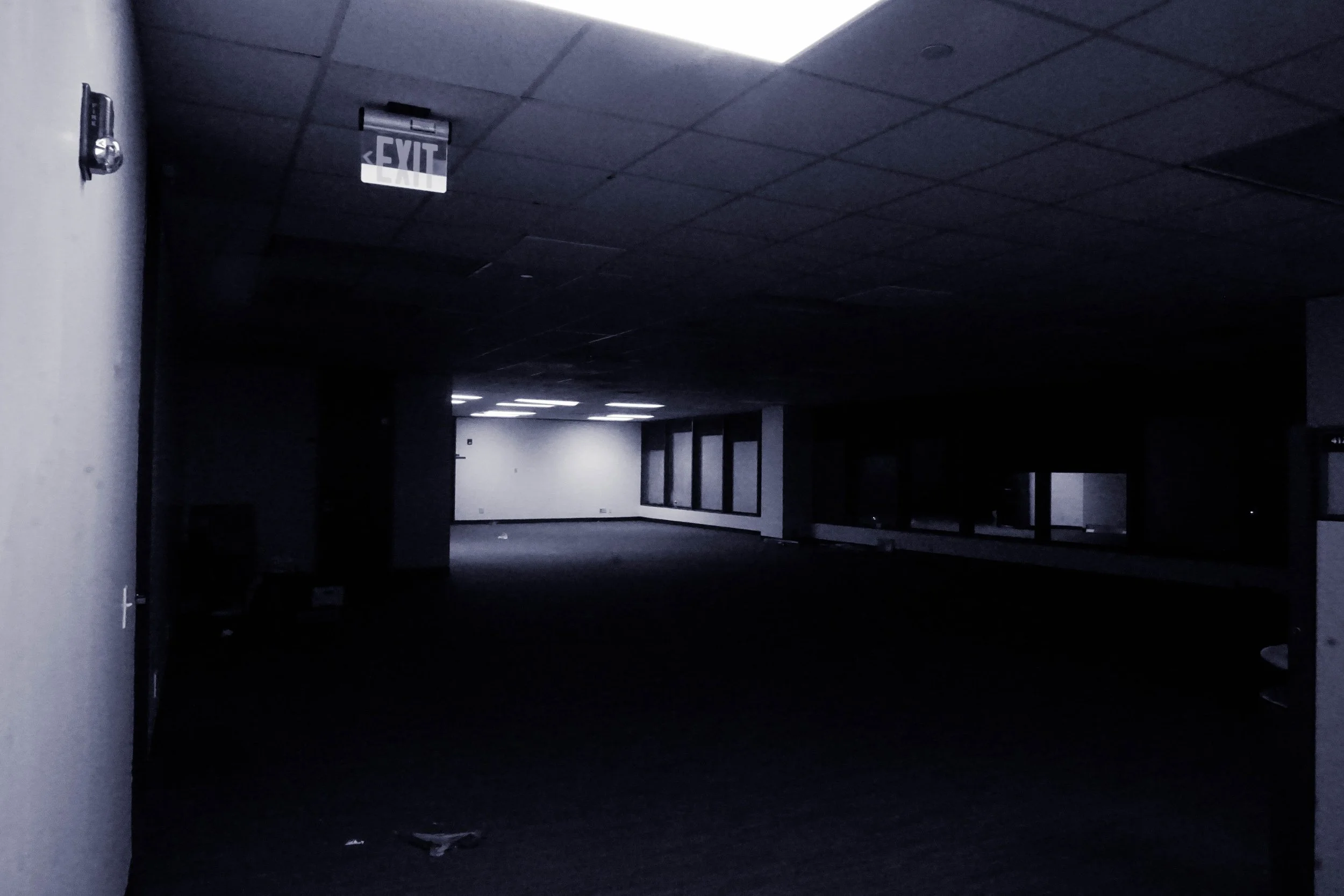 Dark, empty commercial or office space with dim lighting and a visible exit sign on the ceiling.