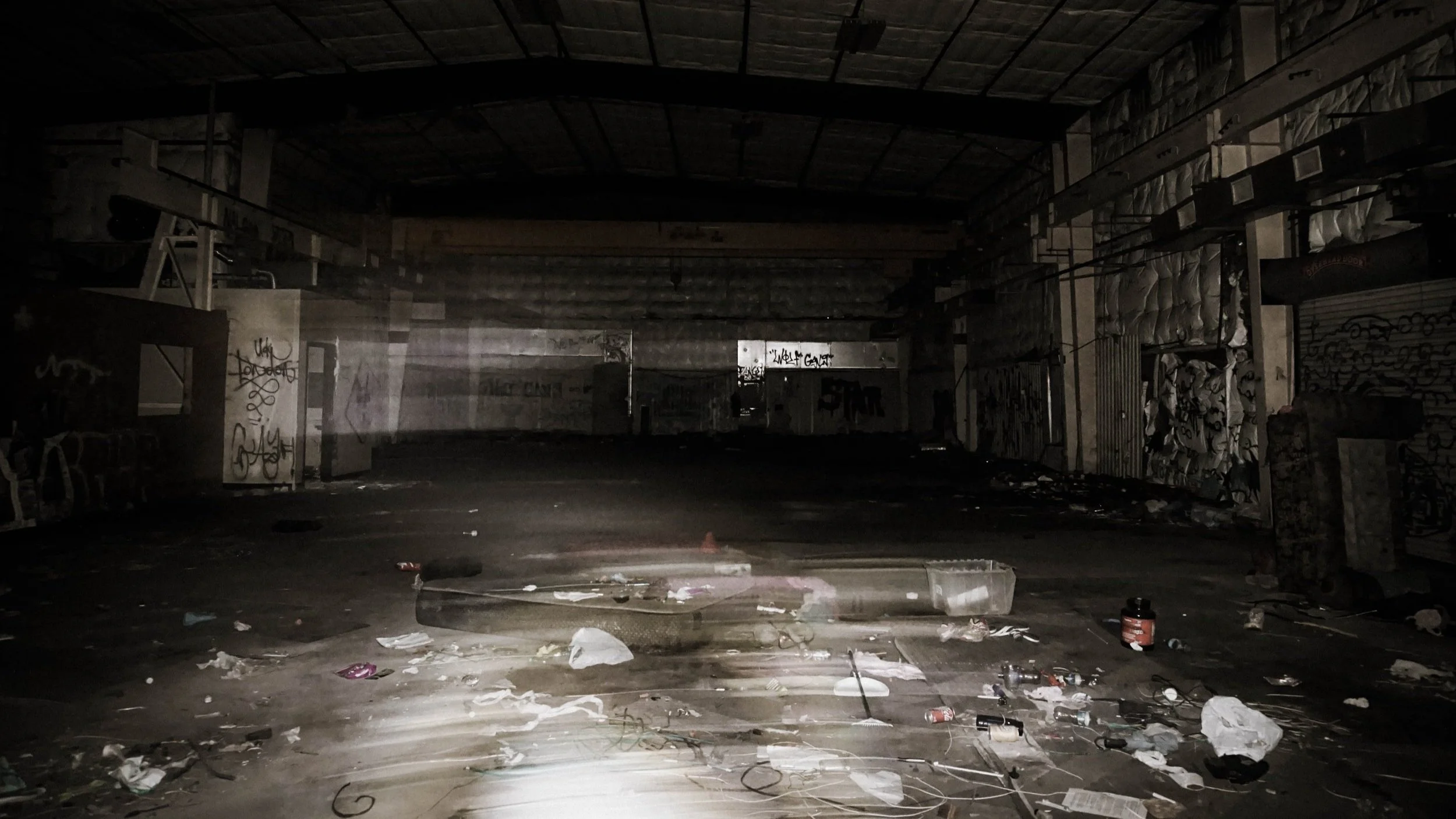 Dark, abandoned indoor space with graffiti-covered walls and scattered debris on the floor, illuminated faintly by a flashlight or camera flash.