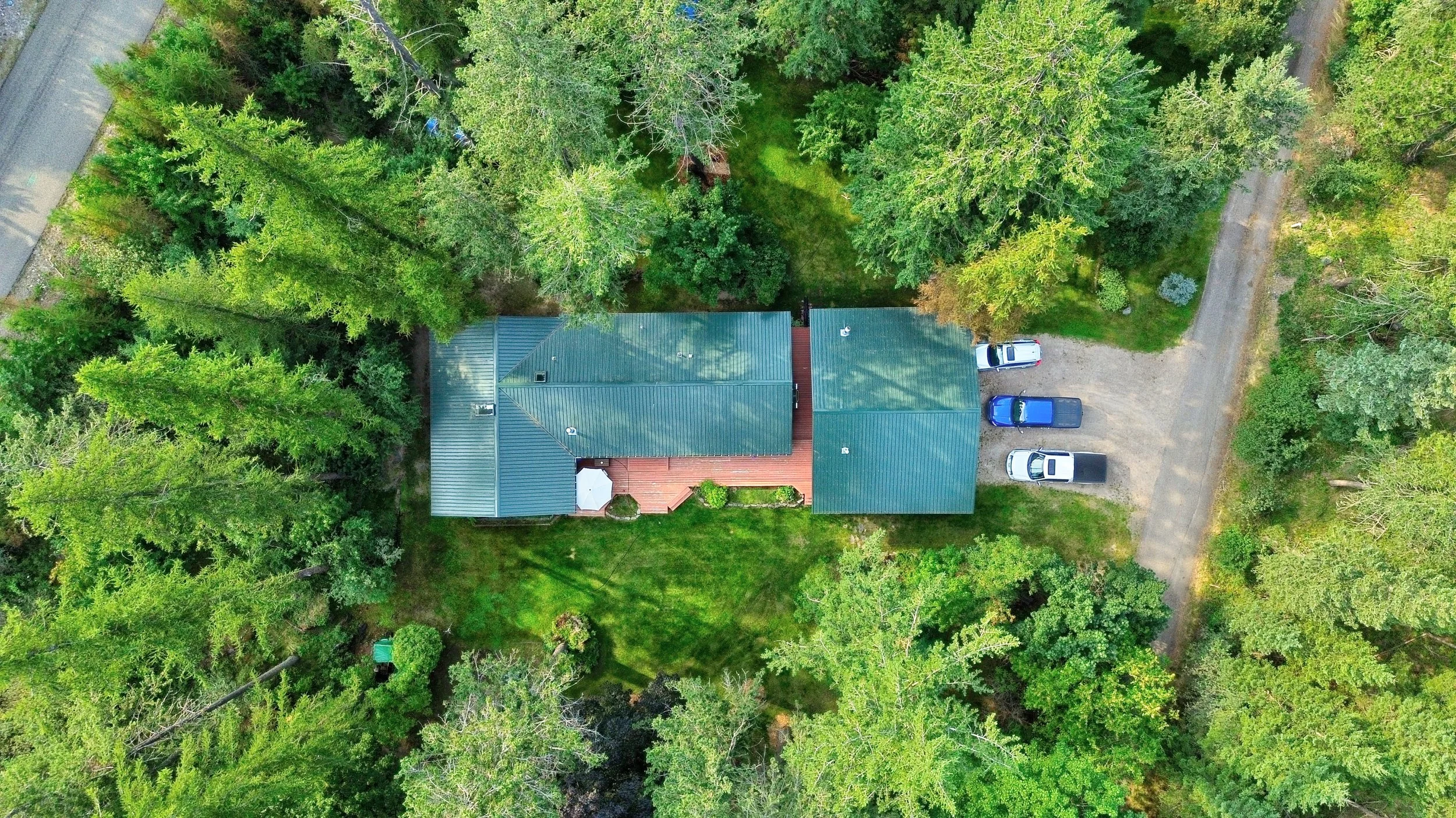 An aerial view of a house with a green roof surrounded by dense green trees, a small deck area with an umbrella, and a gravel driveway with three parked cars.