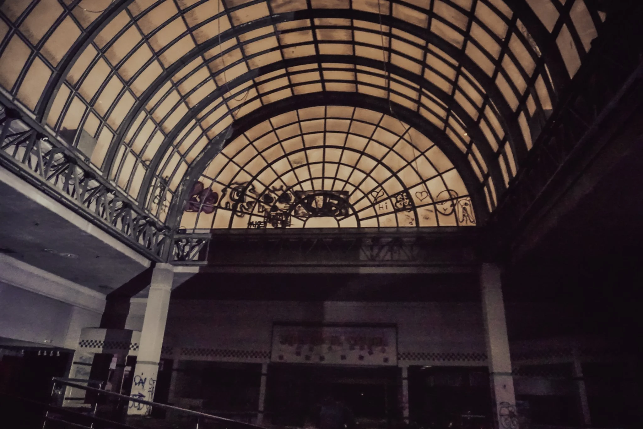 Empty indoor space with a large arched glass ceiling with metal framing, graffiti on the walls, and darkened surroundings.